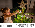 A girl hangs an ornament near the top of a glowing Christmas tree. Decorating the branches lets a child take part in New Year preparations. 143874157