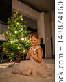 A smiling girl in a tulle dress poses beside a glowing Christmas tree. A holiday portrait celebrates childhood and seasonal traditions at home. 143874160