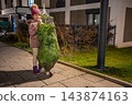 A girl stands beside a net wrapped Christmas tree outside a residential building. Bringing a real tree home begins the next stage of holiday preparations. 143874163