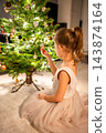 A girl in a pale tulle dress sits beside a lit Christmas tree and reaches toward a bauble. Exploring the ornaments creates a quiet holiday moment at home. 143874164