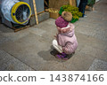 A girl in a winter coat crouches beside a seasonal market stall. A pause during holiday shopping captures childhood curiosity in a public retail space. 143874166
