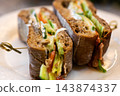 Toasted whole grain sandwich with turkey, avocado, lettuce, tomato, and cream cheese, cut in halves and held by small pick on plate with warm golden crust and visible creamy filling 143874337