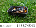 Portable metal barbecue grill with burning charcoal and smoke on green grass field, outdoor picnic preparation 143874543