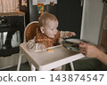 Caring mother hand feeding cute baby boy with spoon in high chair during mealtime at kitchen 143874547