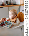 Caring mother hand feeding cute baby boy with spoon in high chair during mealtime at kitchen 143874549