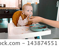Caring mother hand feeding cute baby boy with spoon in high chair during mealtime at kitchen 143874551