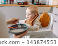 Caring mother hand feeding cute baby boy with spoon in high chair during mealtime at kitchen 143874553