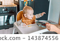 Caring mother hand feeding cute baby boy with spoon in high chair during mealtime at kitchen 143874556