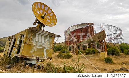 Rusty satellite dish on a wrecked truck in front of the steel frame of an unfinished Soviet solar plant, Armenia 143874827
