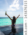 A woman in a blue bikini stands waist deep in an infinity pool, raising a champagne bottle and flute toward a turquoise ocean at a Maldives resort at midday. 143876315