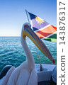 A great white pelican perches on the deck of a boat moving across the ocean beside the national flag of Namibia under a clear daytime sky. 143876317
