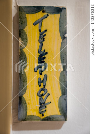 Vertical ceramic plaque reading Telephone hangs on a pale wall in Antibes, France. Soft indoor light reveals glaze, chips, and mottled green ovals framing yellow. Vertical ceramic plaque reading Telephone hangs on a pale wall in Antibes, France. Soft indoor light reveals glaze, chips, and mottled green ovals framing yellow. 143876318
