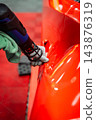 Gloved technician uses a heat gun on a red sports car door handle area inside a luxury car workshop with red floor tiles, focusing on vinyl wrap and surface correction. 143876319
