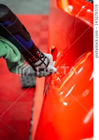 Gloved technician uses a heat gun on a red sports car door handle area inside a luxury car workshop with red floor tiles, focusing on vinyl wrap and surface correction. Gloved technician uses a heat gun on a red sports car door handle area inside a luxury car workshop with red floor tiles, focusing on vinyl wrap and surface correction. 143876319
