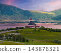 Modern white church with steep roof and square bell tower stands on a grassy hill by a calm fjord in Lofoten, Norway, evening light with pink water reflections. 143876321