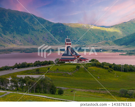 Modern white church with steep roof and square bell tower stands on a grassy hill by a calm fjord in Lofoten, Norway, evening light with pink water reflections. 143876321
