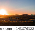 Three travelers stand by a white 4x4 on a dirt track in Namibia. Golden light outlines dry grass, dunes, and rolling hills, wide view, warm gradient, day ending. 143876322