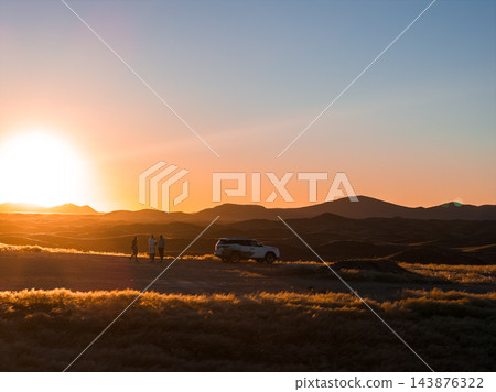 Three travelers stand by a white 4x4 on a dirt track in Namibia. Golden light outlines dry grass, dunes, and rolling hills, wide view, warm gradient, day ending. 143876322