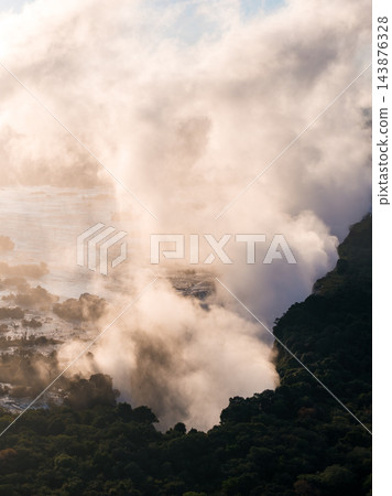 Aerial view shows Victoria Falls at sunset as mist surges above the gorge on the Zimbabwe Zambia border, warm light illuminates spray, dark rainforest lines the cliff edge. 143876328