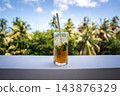 A glass containing a refreshing passion fruit cocktail with mint and ice sits on a white railing, framed by palm trees under a sunny sky in Dhigurah, Maldives. 143876329