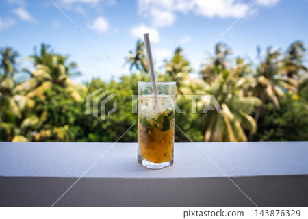 A glass containing a refreshing passion fruit cocktail with mint and ice sits on a white railing, framed by palm trees under a sunny sky in Dhigurah, Maldives. 143876329