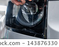 Worker gently aligns a transparent polyurethane protective film onto a vehicle headlight to prevent scratches and stone chips in an indoor detailing garage. 143876330