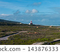 Cape Agulhas Lighthouse stands by the Southern Ocean in South Africa. Fynbos covers flat terrain, a dirt road leads to the beacon, daylight clarity suggests remoteness. 143876333