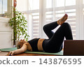 Side view of a mature woman practicing yoga on a mat at home while watching an online fitness lesson on a laptop. Digital wellness and remote training concept. 143876595