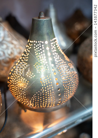 Teardrop shaped perforated metal table lamp glowing warmly among decorative objects. Accent lighting home decor and Egyptian local trade Teardrop shaped perforated metal table lamp glowing warmly among decorative objects. Accent lighting home decor and Egyptian local trade 143877342