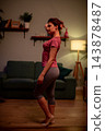 Woman posing in living room, profile view with hand at forehead, subtle stretch and balance near couch and lamp, soft warm lighting, casual workout attire, focused serene routine 143878487