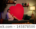 Woman holding large red heart at home. Woman in sweater hugging big red heart on sofa. Relaxed woman in knit sweater clasping giant red heart in cozy living room environment 143878500