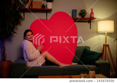 Woman holding large red heart at home. Woman in sweater hugging big red heart on sofa. Relaxed woman in knit sweater clasping giant red heart in cozy living room environment Woman holding large red heart at home. Woman in sweater hugging big red heart on sofa. Relaxed woman in knit sweater clasping giant red heart in cozy living room environment 143878500