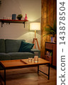 Tall lamp casting warm glow accenting midcentury sofa, plant silhouette, textured wall, curated shelf accessories, atmospheric hotelstyle mood for commercial imagery 143878504