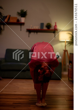 Gentle stretch at home. Maintaining comfort with calming evening stretch routine. Engaging in soothing selfcare stretch on wooden flooring by sofa in warm light Gentle stretch at home. Maintaining comfort with calming evening stretch routine. Engaging in soothing selfcare stretch on wooden flooring by sofa in warm light 143878511