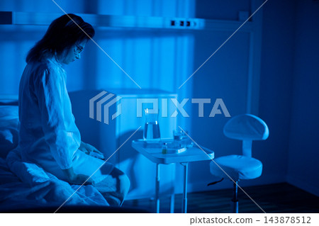 Patient sits on bed near window, pajamas wrinkled, lamp casting soft blue glow, hands folded while staring at distant lights, quiet moment of healing and reflection Patient sits on bed near window, pajamas wrinkled, lamp casting soft blue glow, hands folded while staring at distant lights, quiet moment of healing and reflection 143878512
