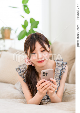一位年輕女子麵朝下躺在家裡的沙發上，正在操作她的智慧型手機。 143881738