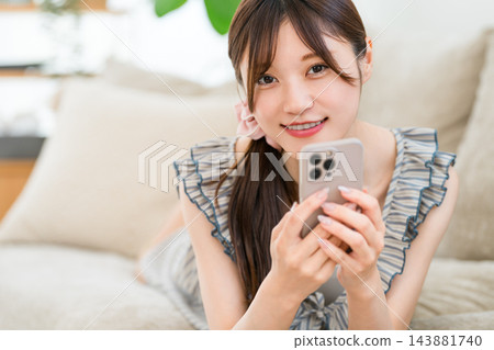 一位年輕女子躺在沙發上，面帶微笑，手裡拿著她的智慧型手機。 143881740