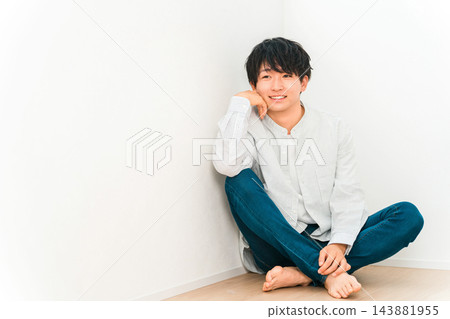 一位年輕男子盤腿而坐，微笑地倚靠在白牆上。 143881955
