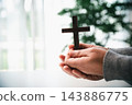 Two hands in gray sleeves hold a plain wooden cross upright near a bright window, with softly blurred greenery behind it in a vertical close-up suited to Christian faith content. 143886775