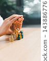 Two hands form a protective gesture around wooden peg figures and stacks of coins on a desk, creating a vertical concept image about caring for people and planning financial resources. 143886776