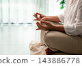 A person sits cross-legged indoors with hands resting on the knees in a meditation-style gesture beside a bright window, leaving wide space on the left for mindfulness and wellness text. 143886778