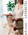 A cropped vertical view shows a woman sitting cross-legged on a floor cushion with open palms resting on her knees, beside a bright window and leafy plant in a softly lit room at home. 143886781