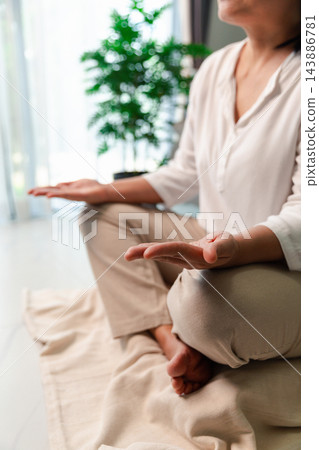 A cropped vertical view shows a woman sitting cross-legged on a floor cushion with open palms resting on her knees, beside a bright window and leafy plant in a softly lit room at home. 143886781