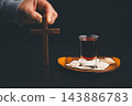 Hand holding a wooden cross beside communion wafers and dark red beverage on a wooden plate, representing Christian worship, faith, prayer, sacred remembrance, devotion, 143886783