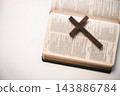 A plain wooden cross rests diagonally across the pages of an open book on a white surface, leaving wide copy space on the left for Christian faith or reflection text. 143886784