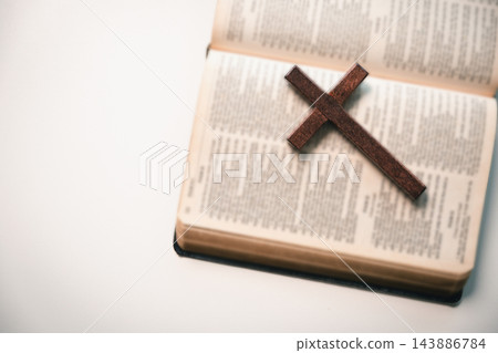 A plain wooden cross rests diagonally across the pages of an open book on a white surface, leaving wide copy space on the left for Christian faith or reflection text. A plain wooden cross rests diagonally across the pages of an open book on a white surface, leaving wide copy space on the left for Christian faith or reflection text. 143886784