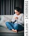A woman wearing a gray cardigan and cross necklace sits with legs folded on a sofa, head bowed and hands clasped near her chest, leaving space on the left for faith or reflection text. 143886802