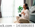 A softly blurred woman sits cross-legged on a floor cushion with both hands near her chest, beside a bright window, potted plant, and sofa, leaving wide copy space on the left for wellness text. 143886803
