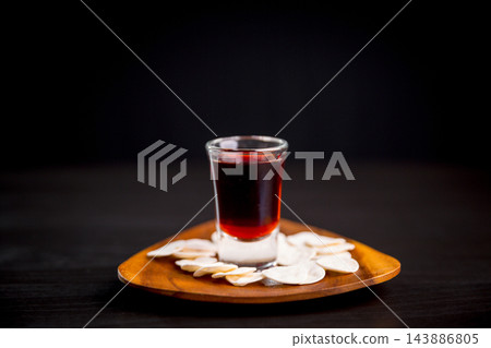 Dark red beverage in a clear glass surrounded by communion wafers on a wooden plate, creating a simple Christian still life for worship, prayer, faith, sacred remembrance, and reflection. Dark red beverage in a clear glass surrounded by communion wafers on a wooden plate, creating a simple Christian still life for worship, prayer, faith, sacred remembrance, and reflection. 143886805