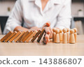A hand stands between a row of leaning wooden domino blocks and several wooden peg figures, illustrating an attempt to interrupt a chain reaction before it reaches the figures. 143886807